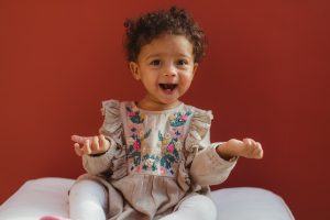 toddler girl in flower dress with red background holding hands up in question