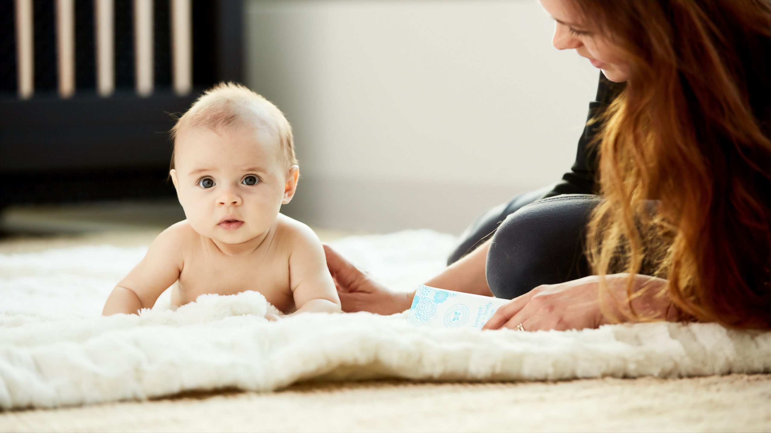 adorable baby on soft rug lying next to mom with crib behind them